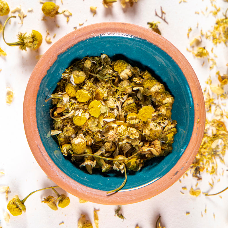 Chamomile herb in blue clay bowl with white background and herb surrounding.