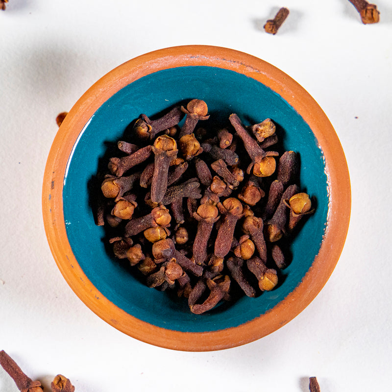 Cloves herb in blue clay bowl with white background and herb surrounding.