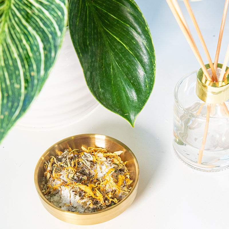 Postpartum Sitz Bath in a gold bowl filled with Epsom Salt, Calendula, Comfrey, Lavender, Plantain, Red Raspberry, Shepherd’s Purse. Surrounded by green plant and glass diffuser.